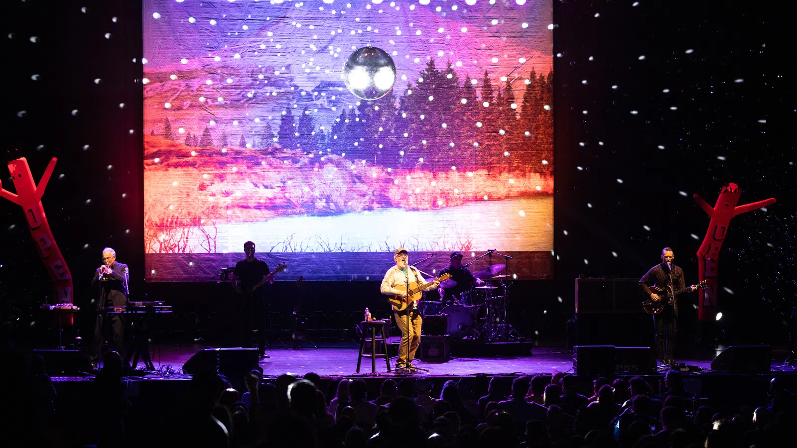Band performing on stage with snow effects and forest landscape backdrop under disco ball lighting.