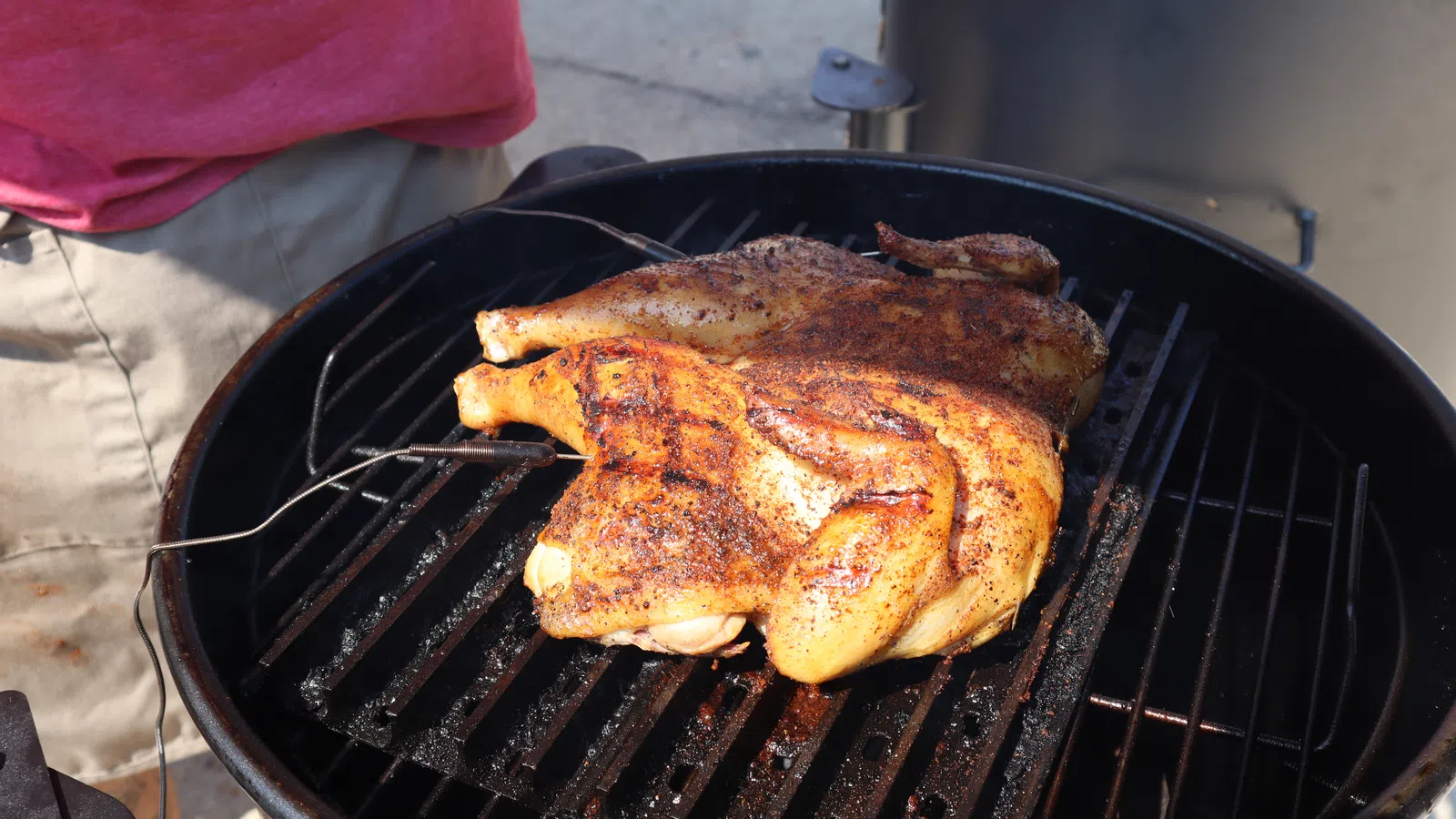 Spatchcock chicken seasoned and grilling on a charcoal barbecue with a probe thermometer inserted.