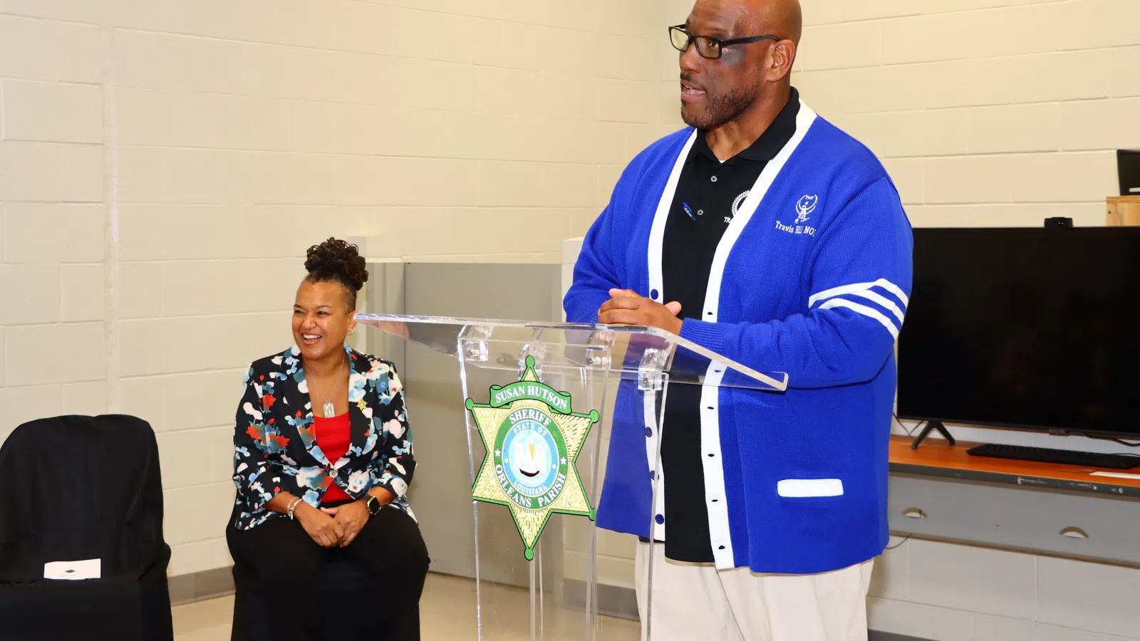Man in blue cardigan speaking at podium with sheriff badge, woman in floral blazer seated nearby smiling