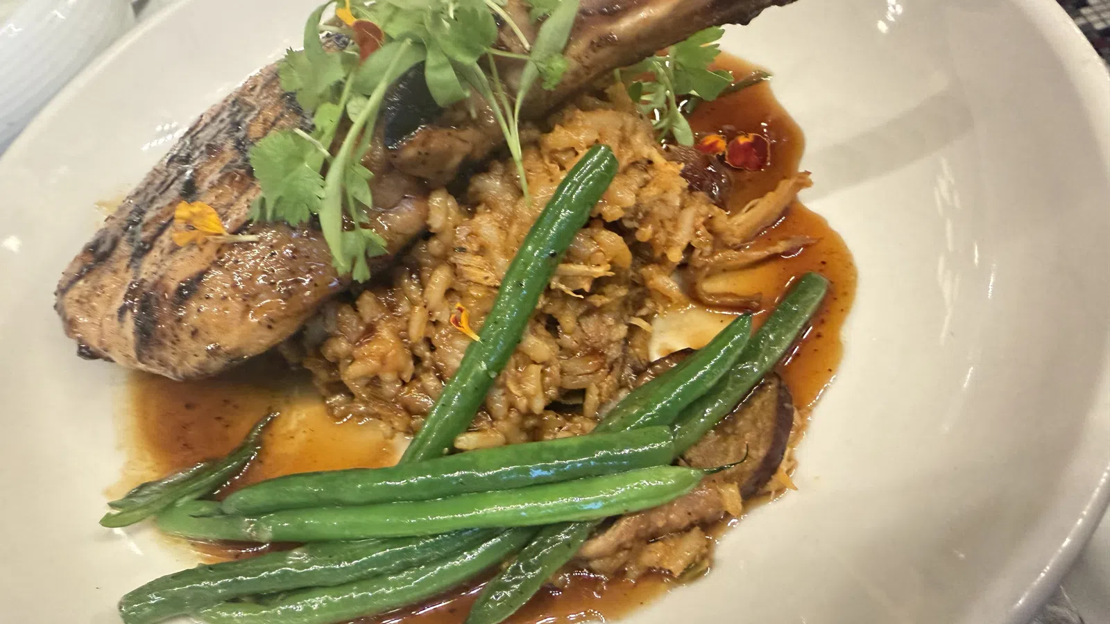 Grilled pork chop with green beans, rice, and sauce garnished with fresh herbs in a white bowl.