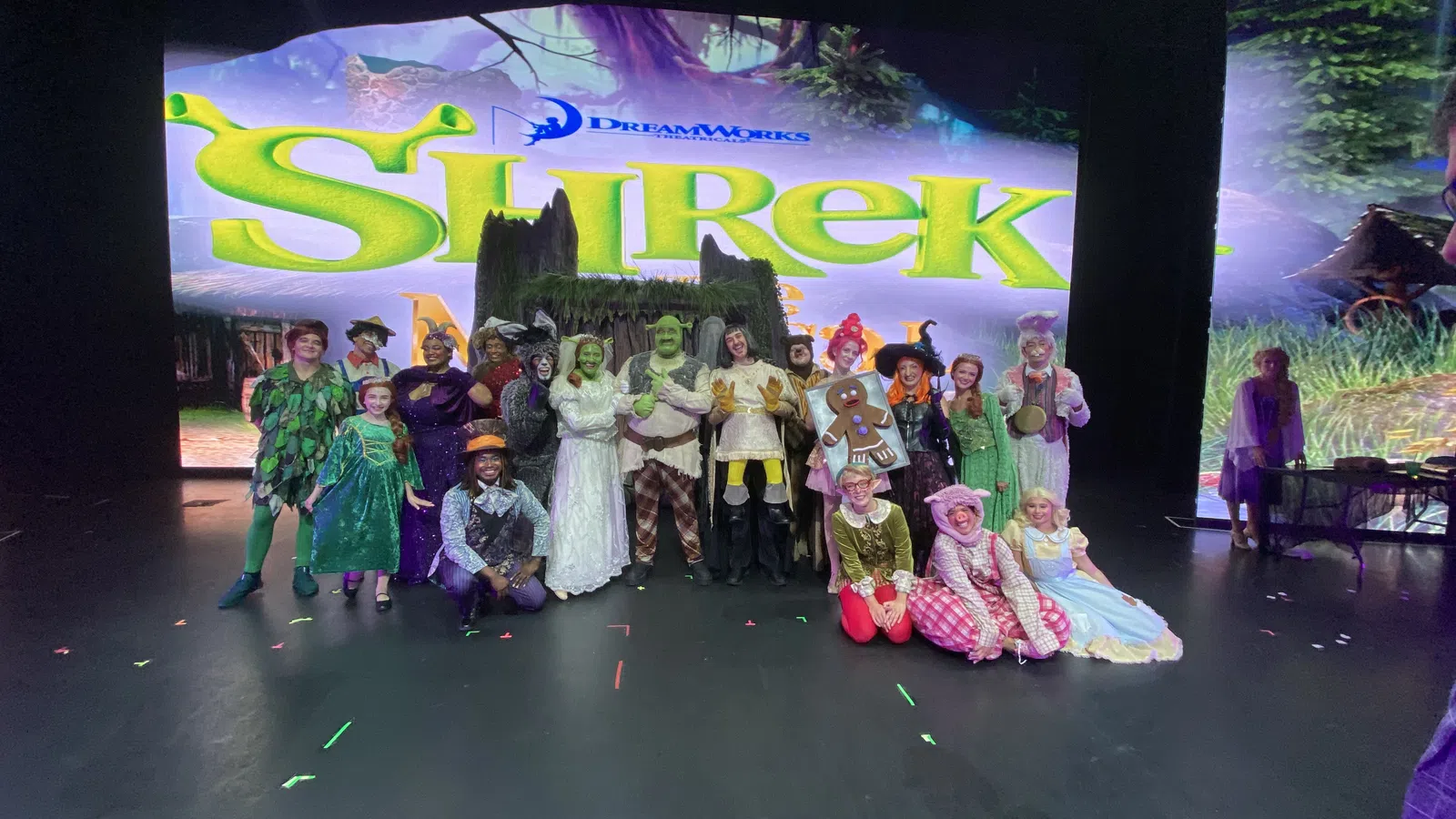 Cast of Shrek musical in full costumes posing on stage with Shrek show title backdrop