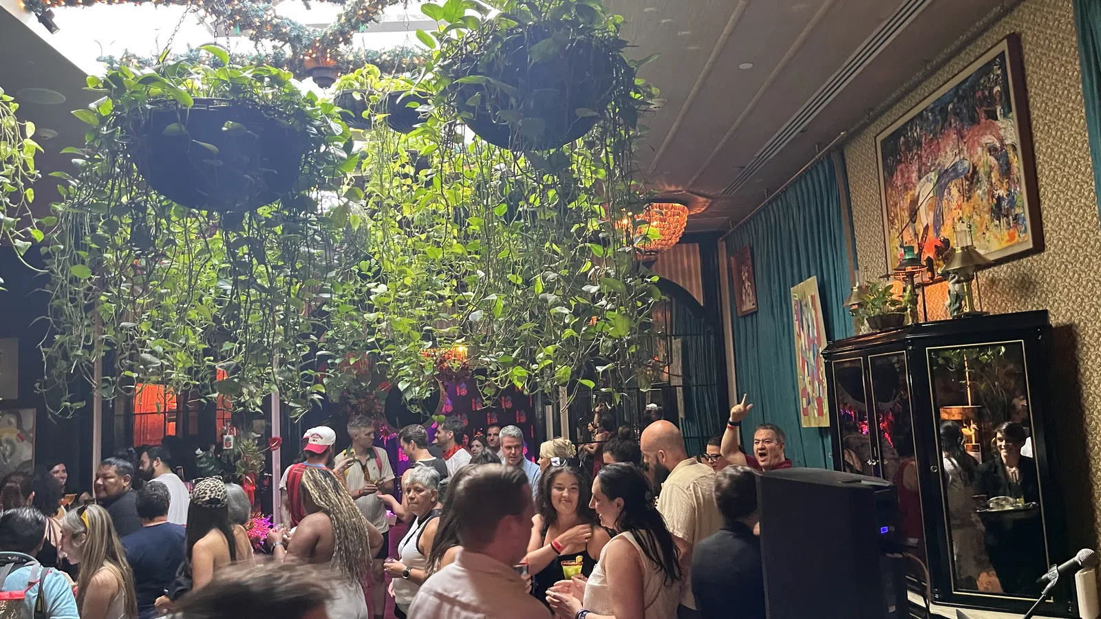 Crowded indoor party with hanging plants, colorful lighting, and guests socializing and enjoying drinks.