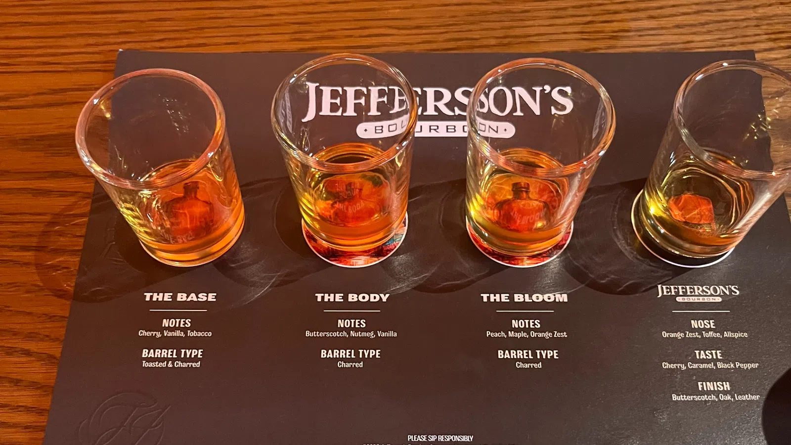 Four whiskey tasting glasses with varying bourbon samples on a Jefferson's Bourbon tasting mat showing flavor notes and barrel types.