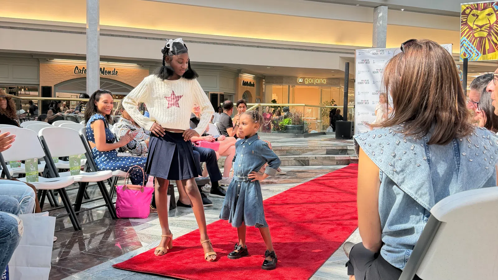 Two young models confidently posing on a red carpet runway during a fashion event in a shopping mall.
