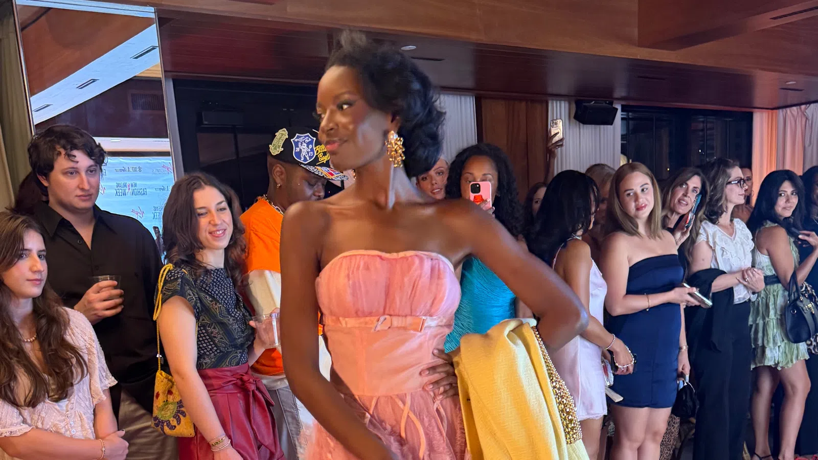 Model in pink gown holding yellow jacket walking past crowd of spectators at indoor event with wood paneling.