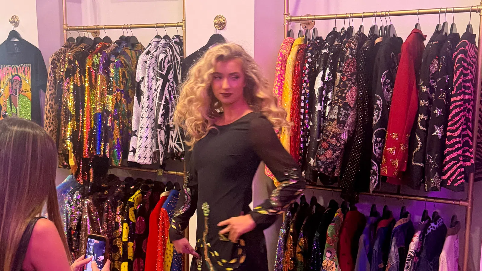 Woman in black dress posing in a colorful boutique with sequined and patterned jackets on racks