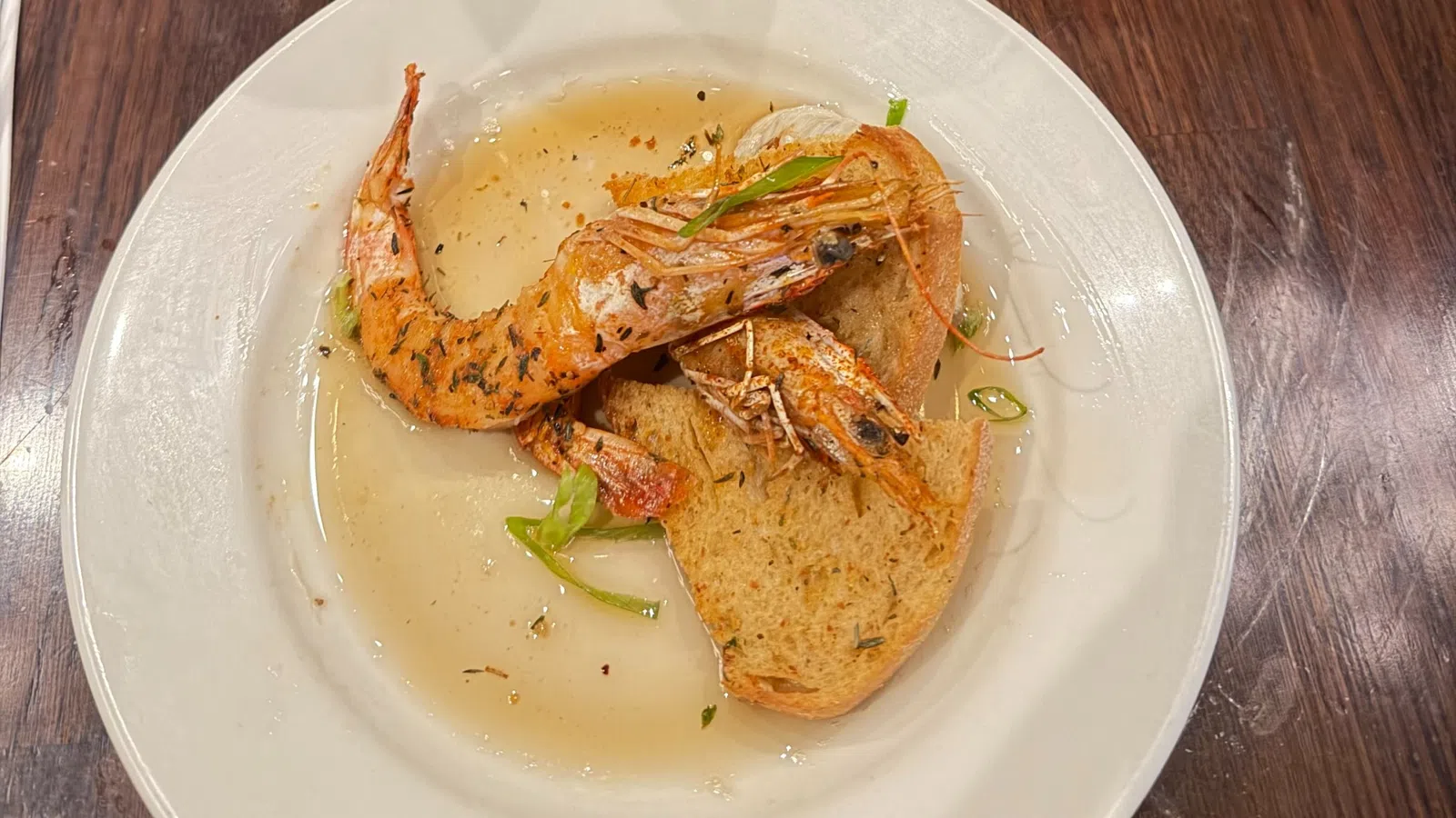 Two grilled shrimp served on toasted bread with broth and garnished with herbs on a white plate.
