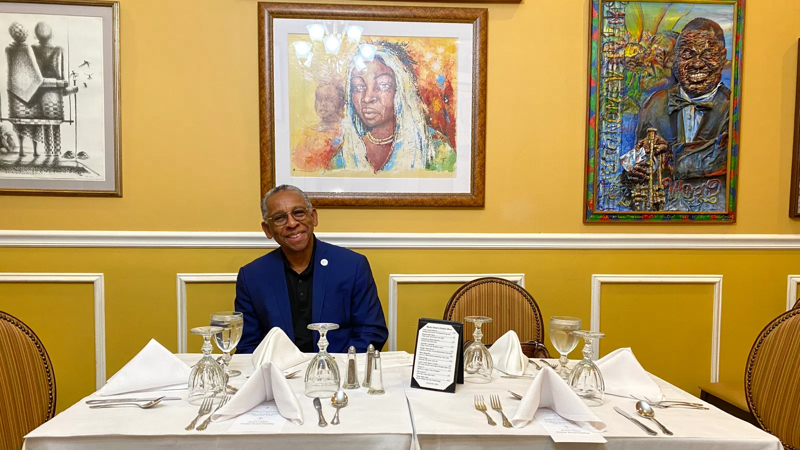 Man in blue suit sitting at white tablecloth-covered table set with wine glasses and silverware, framed art on yellow wall.