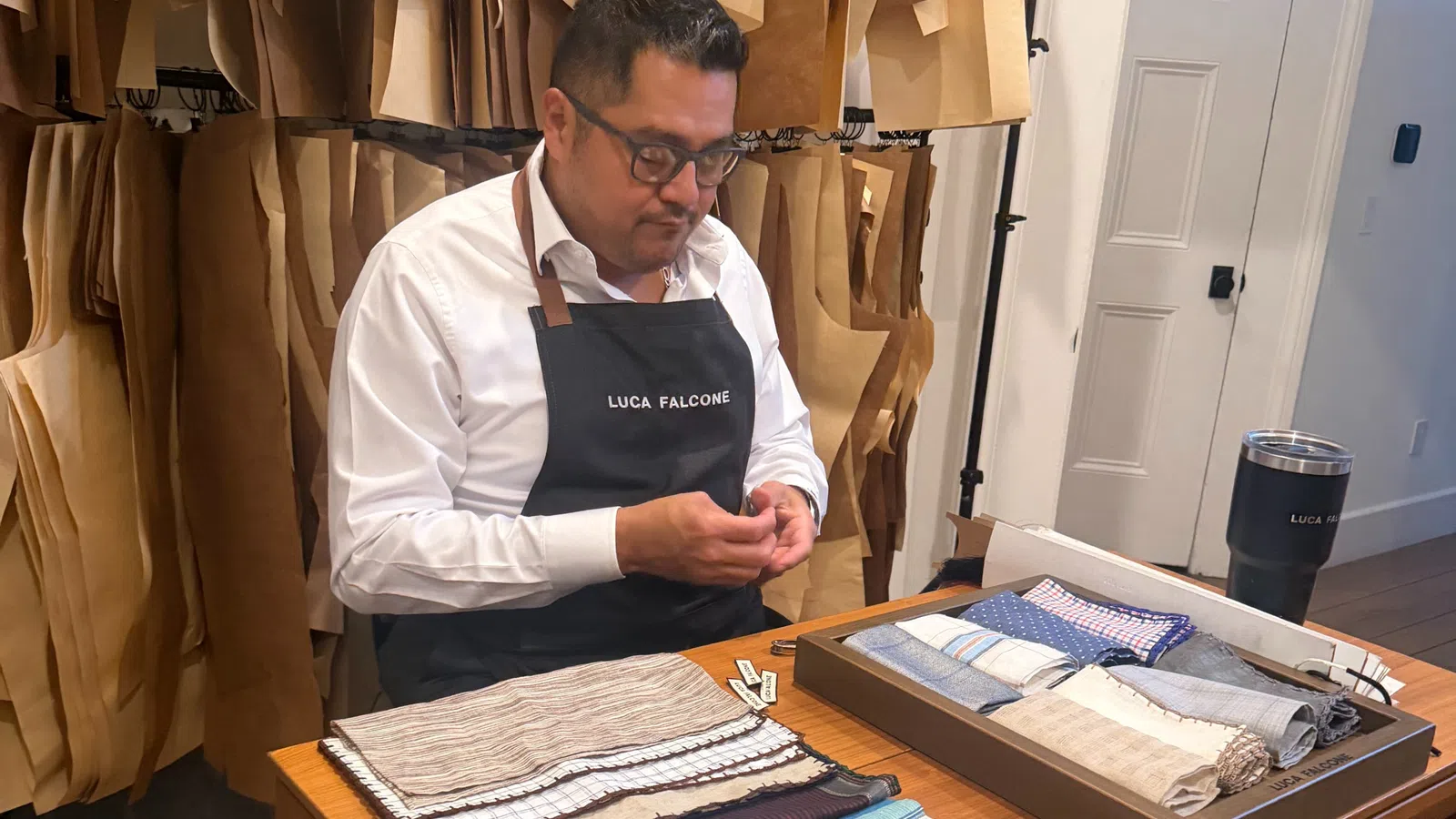 Tailor wearing Luca Falcone apron working with fabric samples in a workshop filled with hanging patterns.