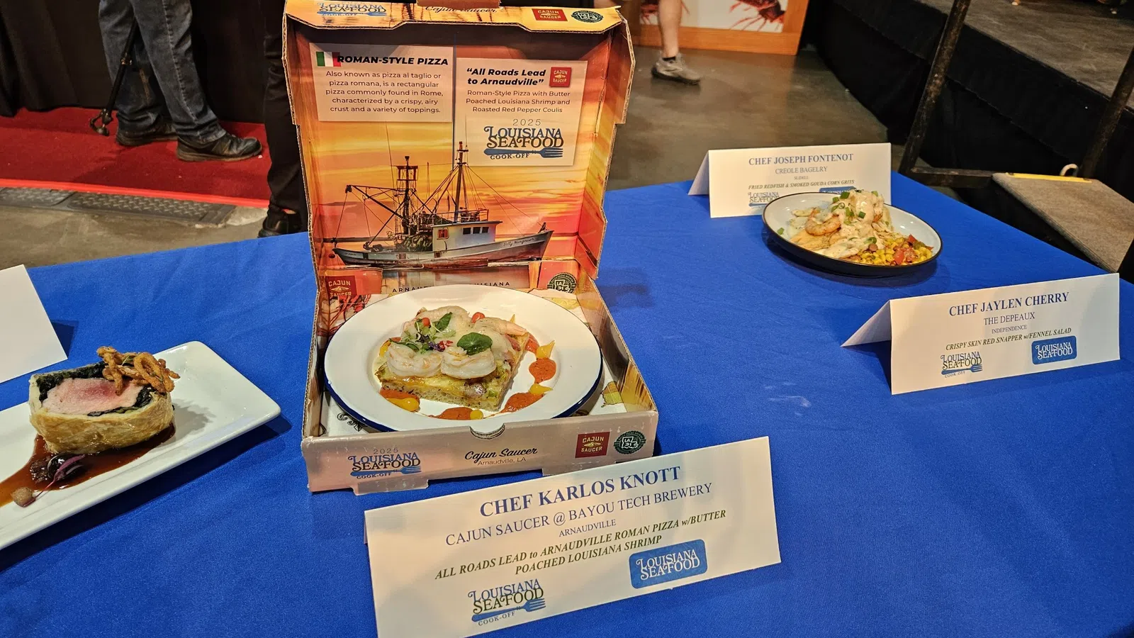 Gourmet seafood dishes displayed on a blue tablecloth with chef name cards at Louisiana Seafood event.
