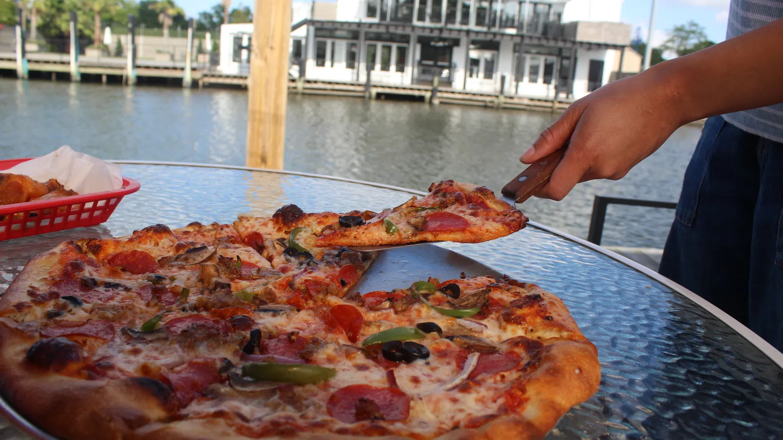 Person lifting a slice of pepperoni pizza from a glass table by the waterfront with a house in the background