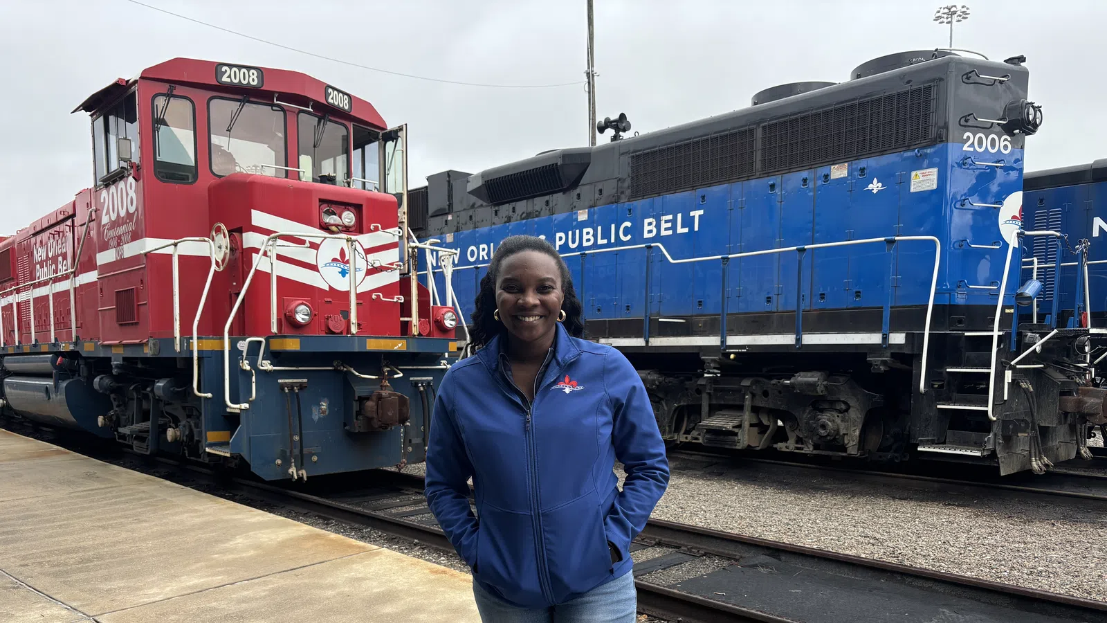 Smiling woman in blue jacket standing on platform between red and blue trains at a railway yard.
