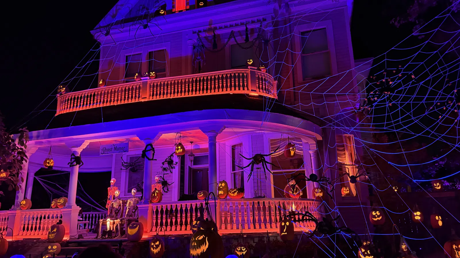 Large Victorian house decorated with glowing jack-o'-lanterns, skeletons, giant spiders, and web lights for Halloween night.