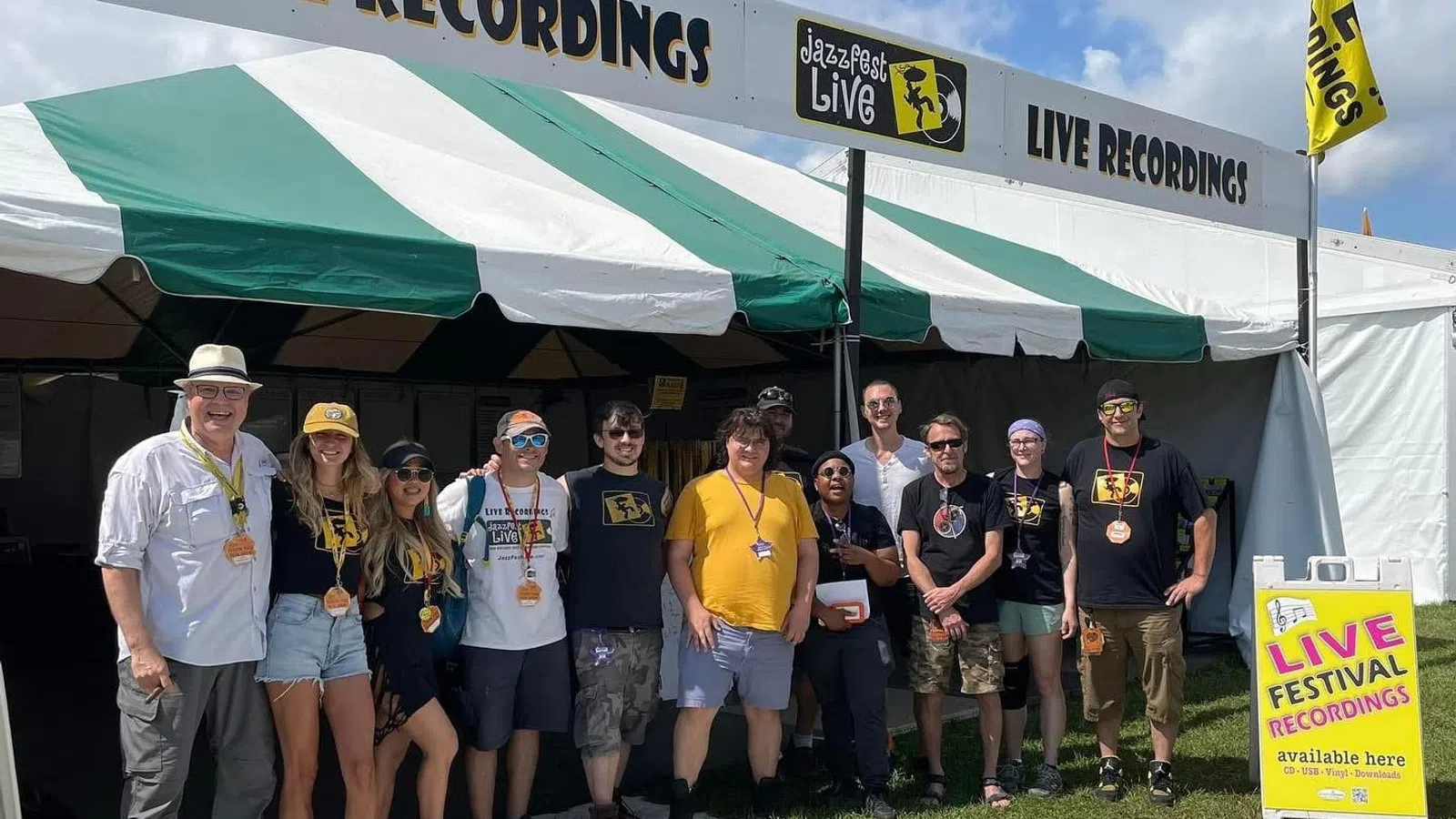 Group of people standing in front of a Jazz Fest Live tent offering live festival recordings during a sunny day.