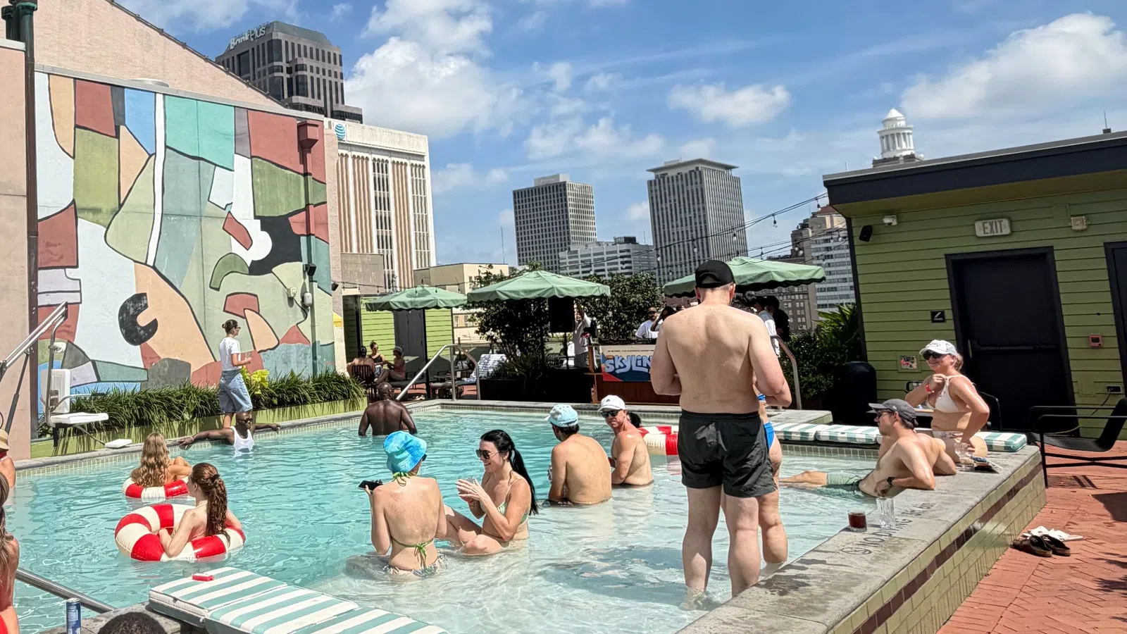 People relaxing and socializing in a rooftop pool with city skyline and colorful mural background on a sunny day.