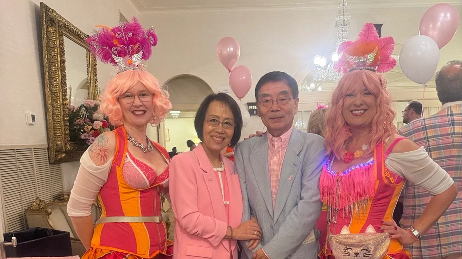 Two women in colorful circus costumes and pink wigs pose with a couple dressed in pastel pink and gray at a festive event.