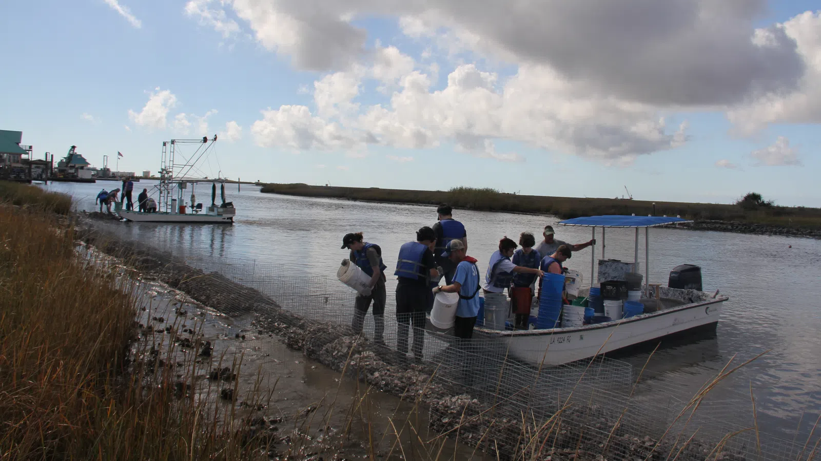 Group of people working on a boat near a marshy shoreline under a cloudy sky during daytime