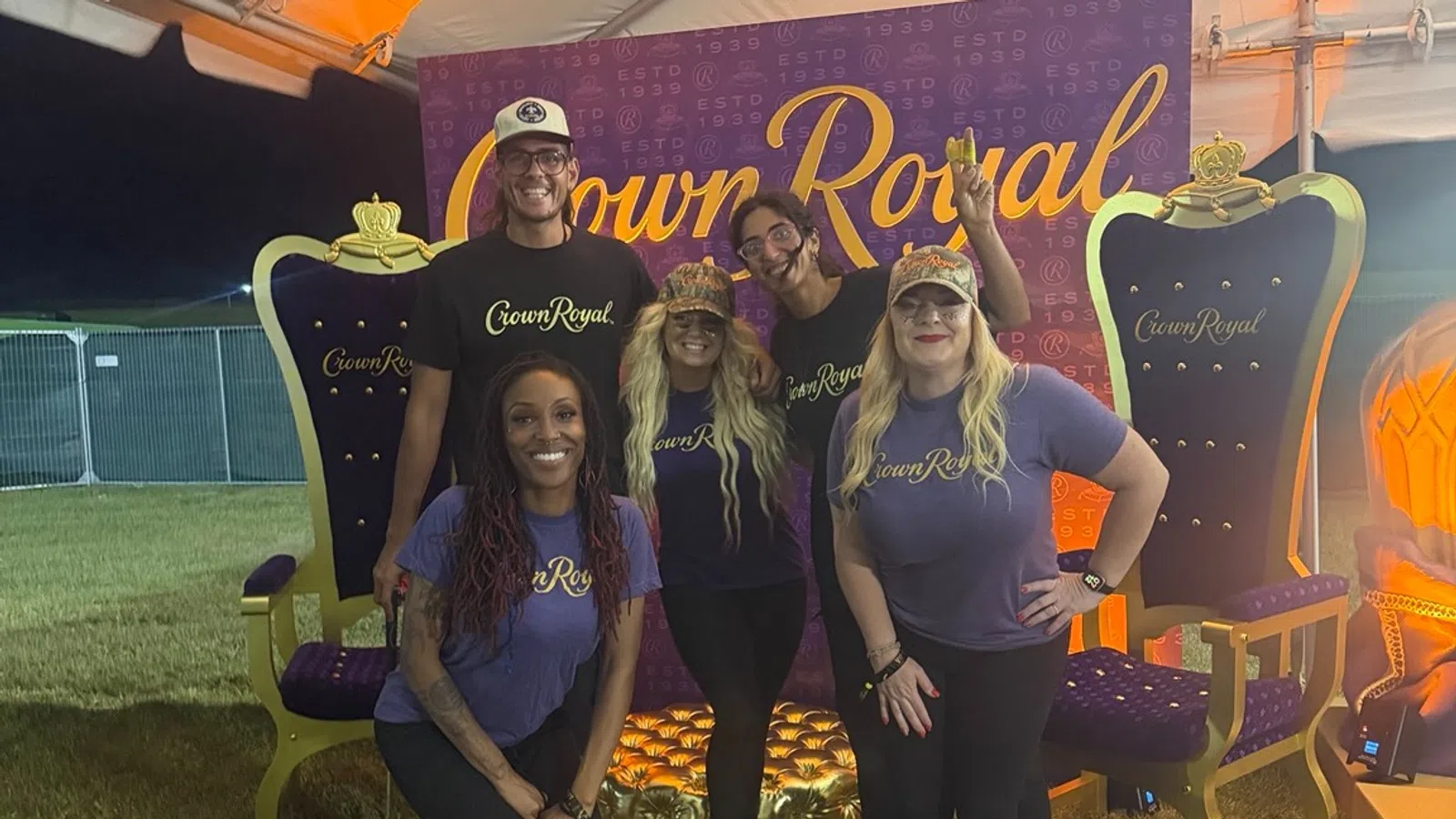 Five people wearing Crown Royal shirts pose in front of a Crown Royal backdrop between two large decorative royal chairs.