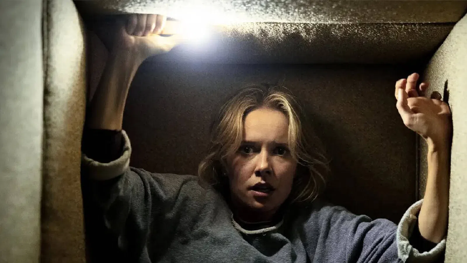 Woman trapped in a small dark space holding a bright flashlight above her head, looking fearful and tense.