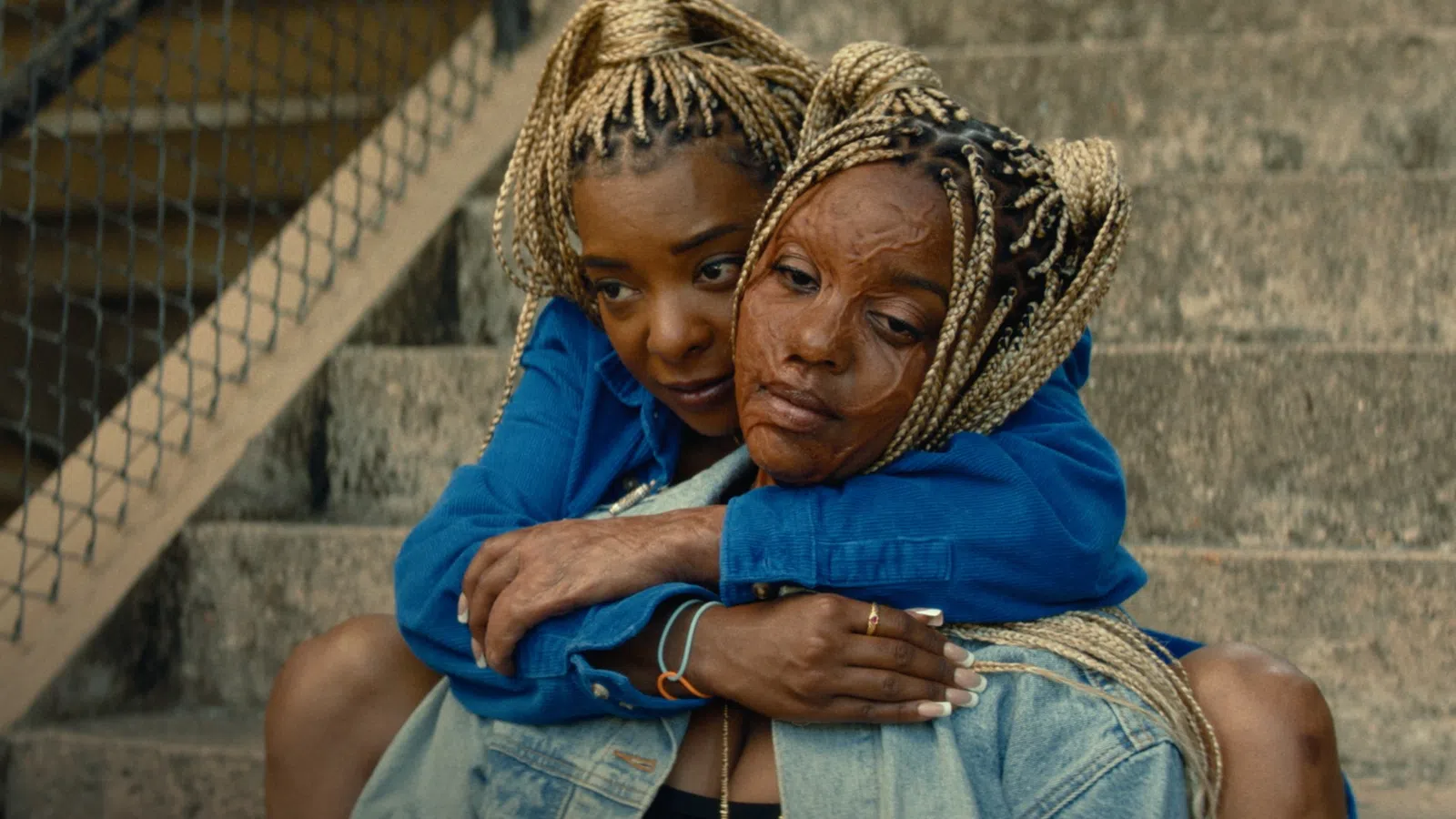 Two women with braided hair embrace on concrete stairs, showing support and emotional connection.