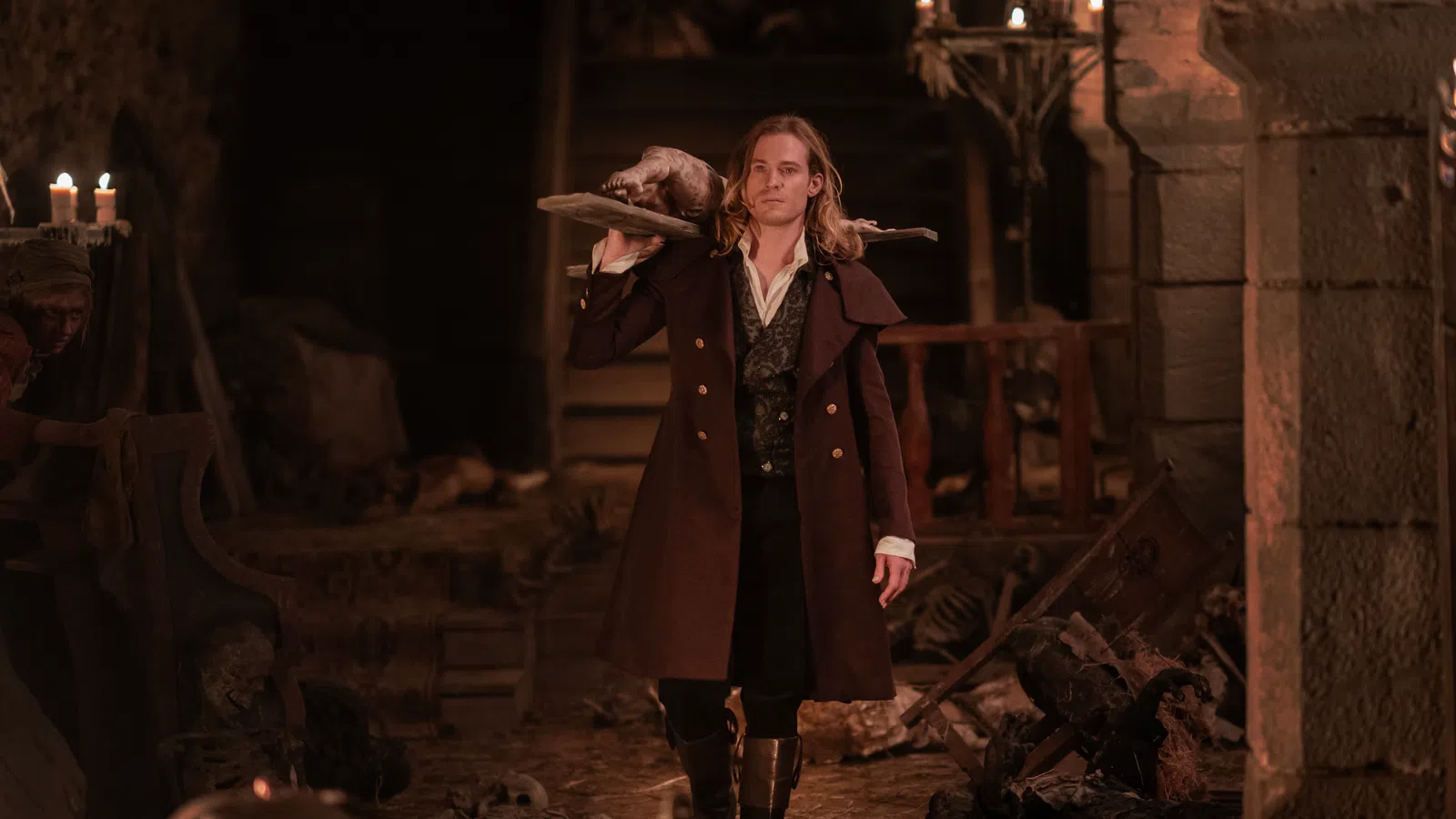 Man in historical coat carrying a wooden board with a dead animal in a dimly lit stone room with skeletons.