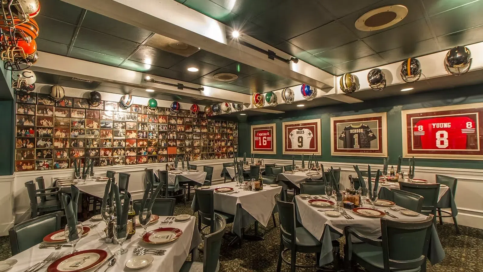 Sports-themed restaurant dining area with framed jerseys, helmets, and photo collage on walls, set tables with plates.