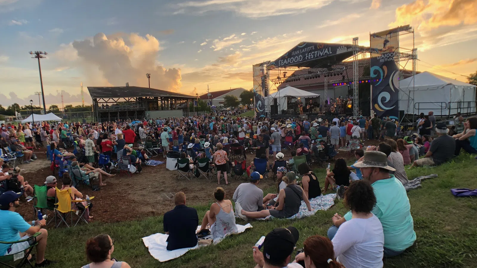 Crowd enjoying live music at Jean Lafitte Seafood Festival during sunset with colorful sky and stage lighting.