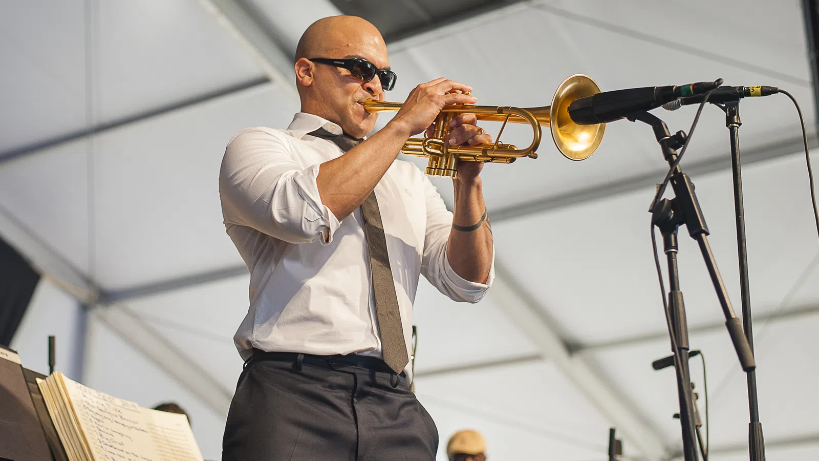 Bald man wearing sunglasses playing trumpet on stage under white tent with microphone nearby