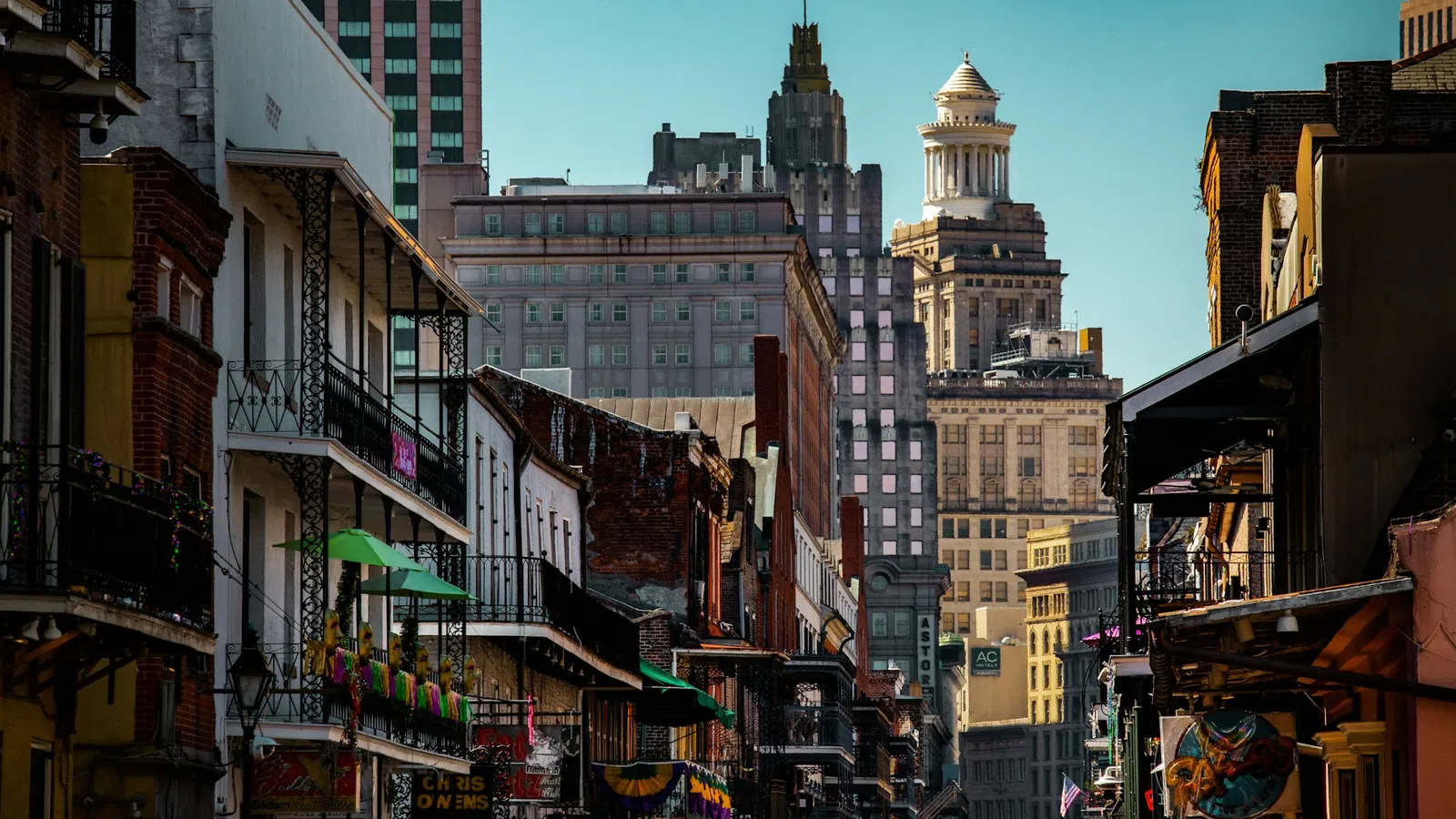 Historic New Orleans street with balconies, colorful decorations, and city skyline in the background under blue sky.