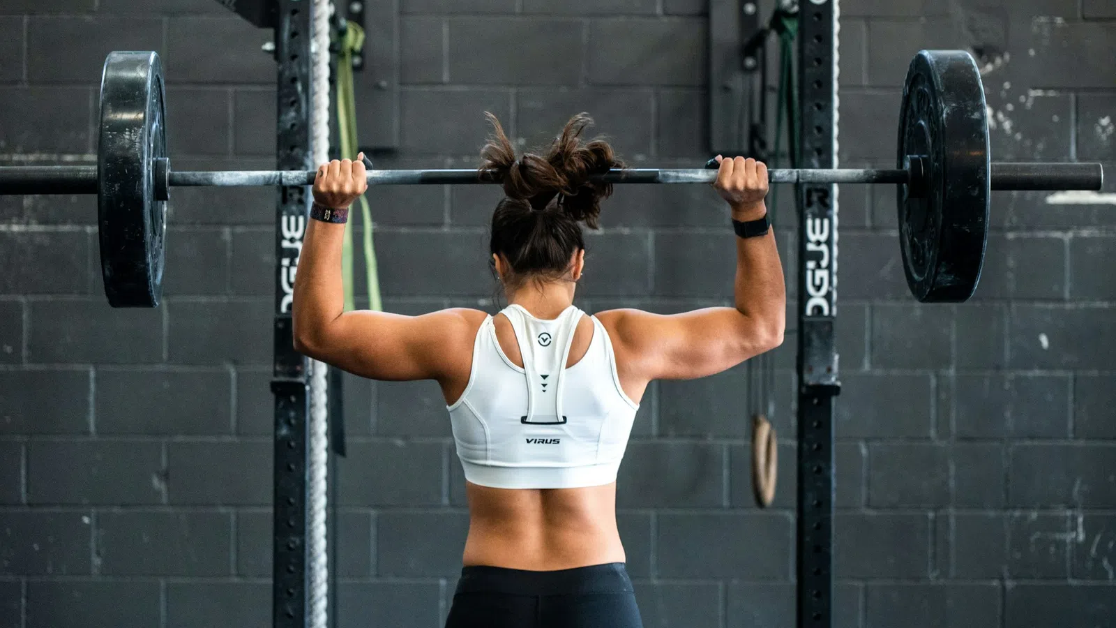 Woman lifting a heavy barbell overhead in a gym with a black brick wall background
