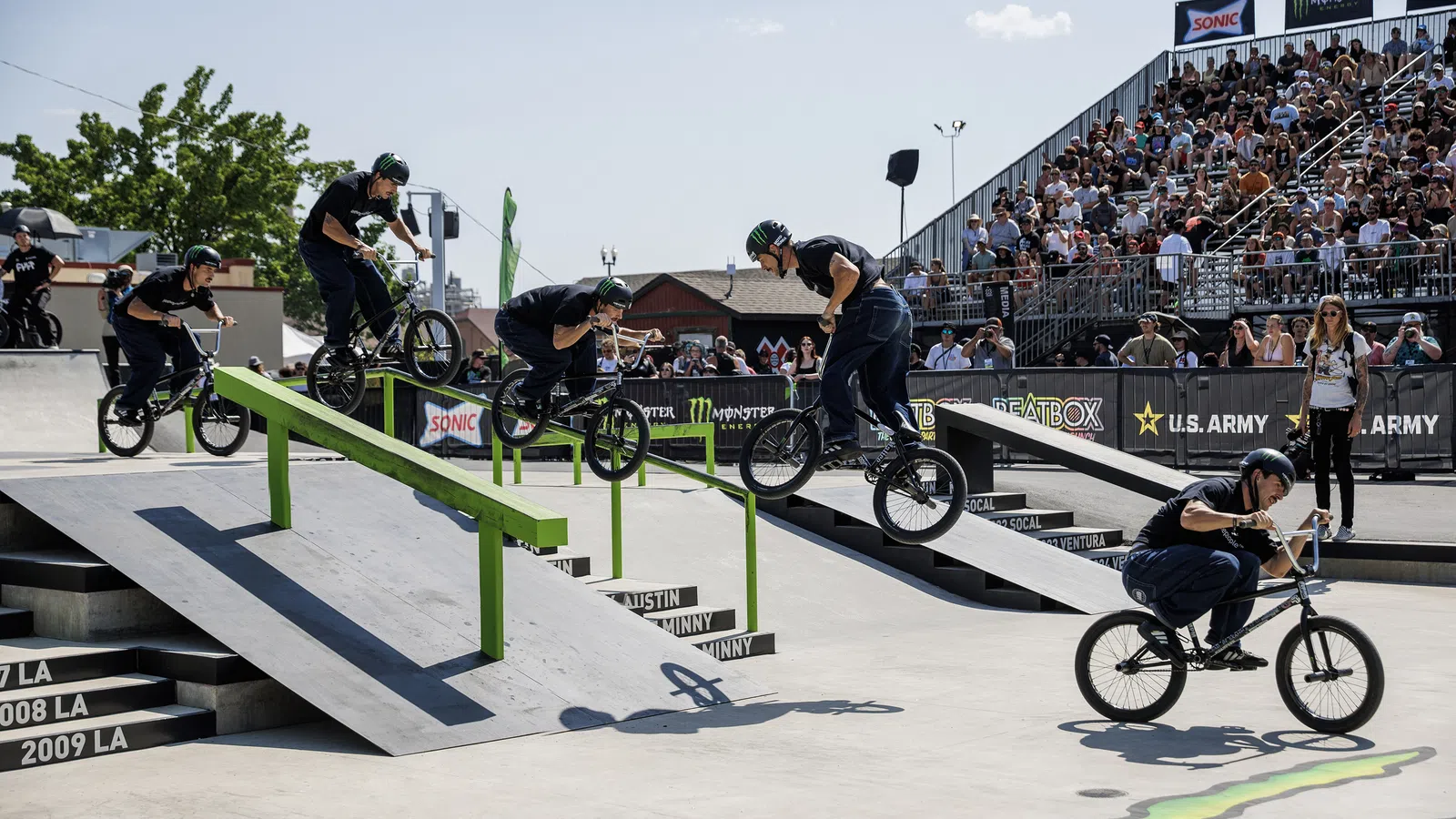 BMX rider performing a stunt sequence over a green rail in a skatepark with a large crowd watching.