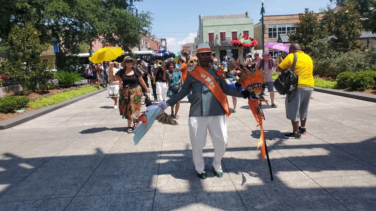 Man wearing a striped suit, orange sash, and hat leads a lively street parade with musicians and colorful umbrellas.