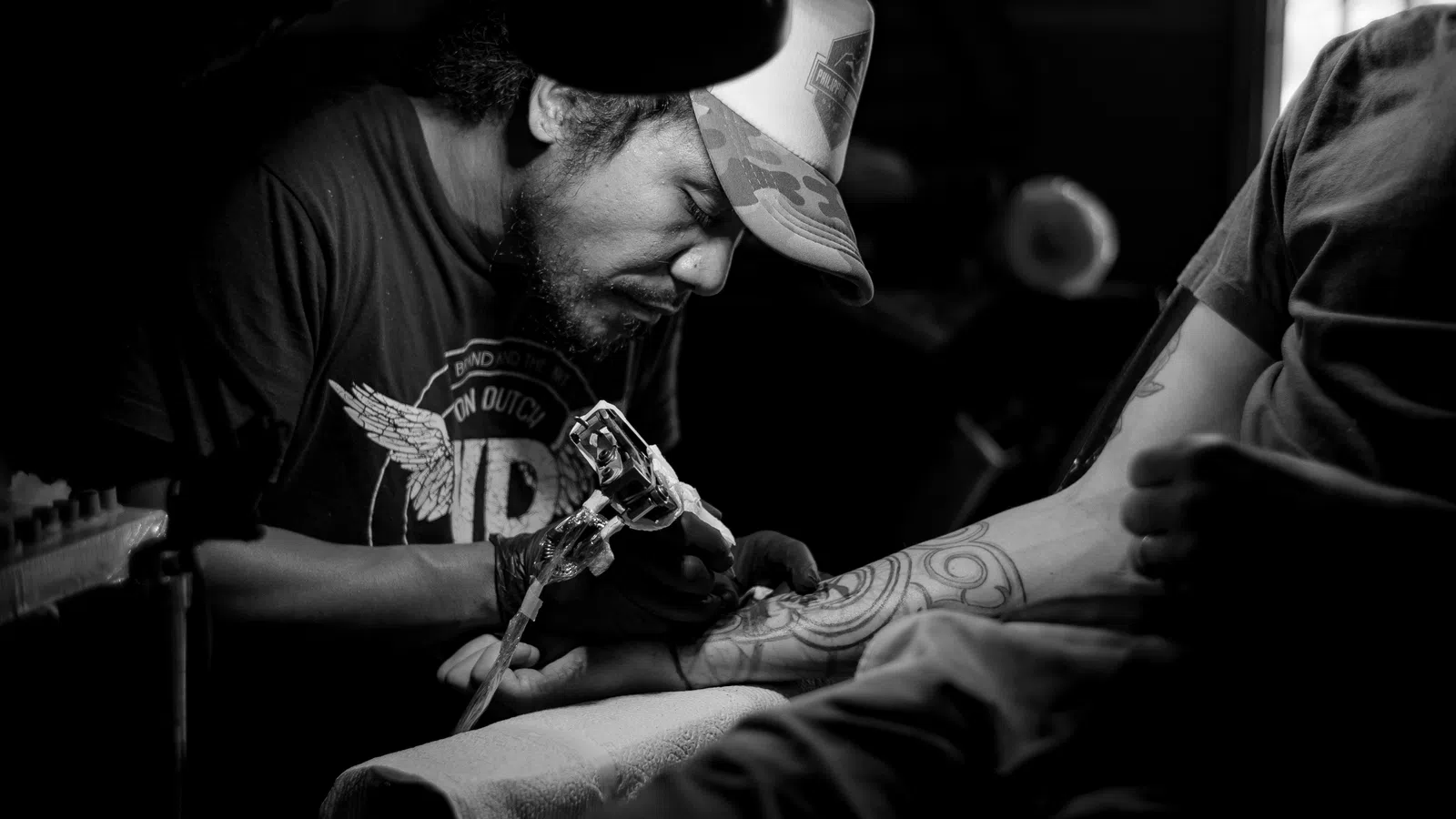 Black and white photo of a tattoo artist focused on inking a geometric tattoo on a client's forearm in dim light.