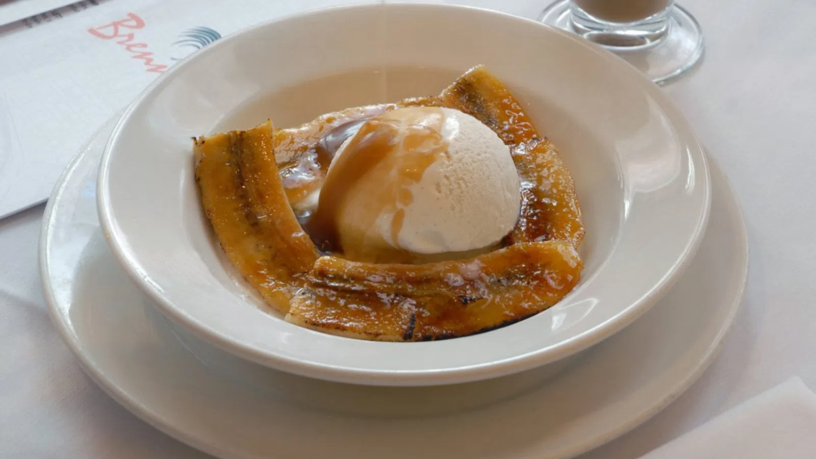 Grilled plantains topped with vanilla ice cream and caramel sauce in a white bowl on a white table.