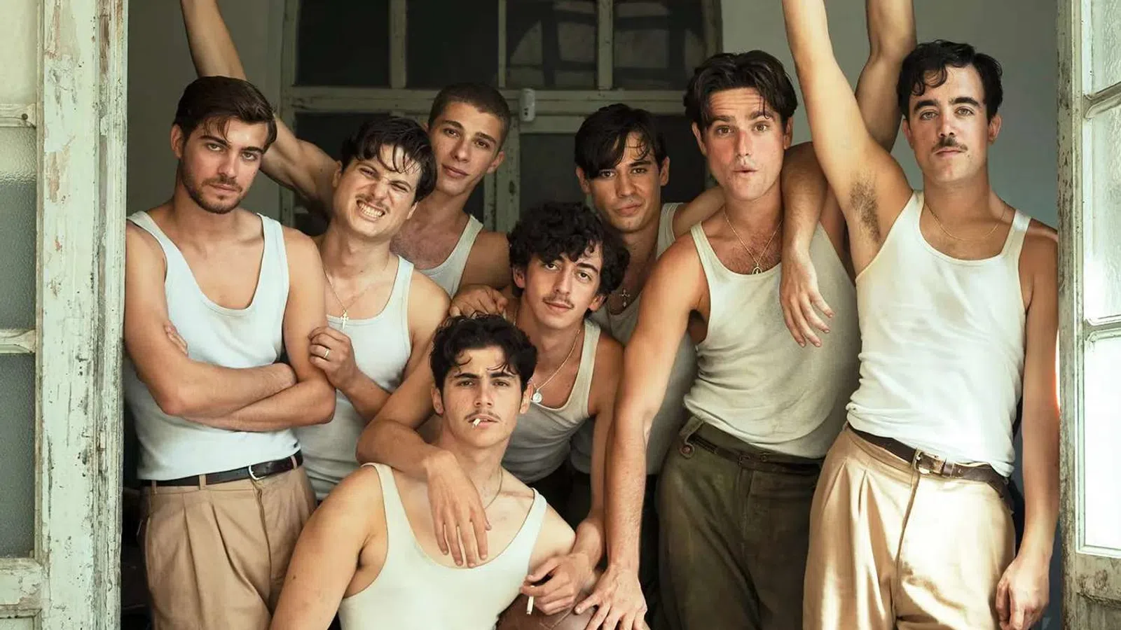 Group of eight young men wearing white tank tops and beige pants posing closely in a vintage room near open windows