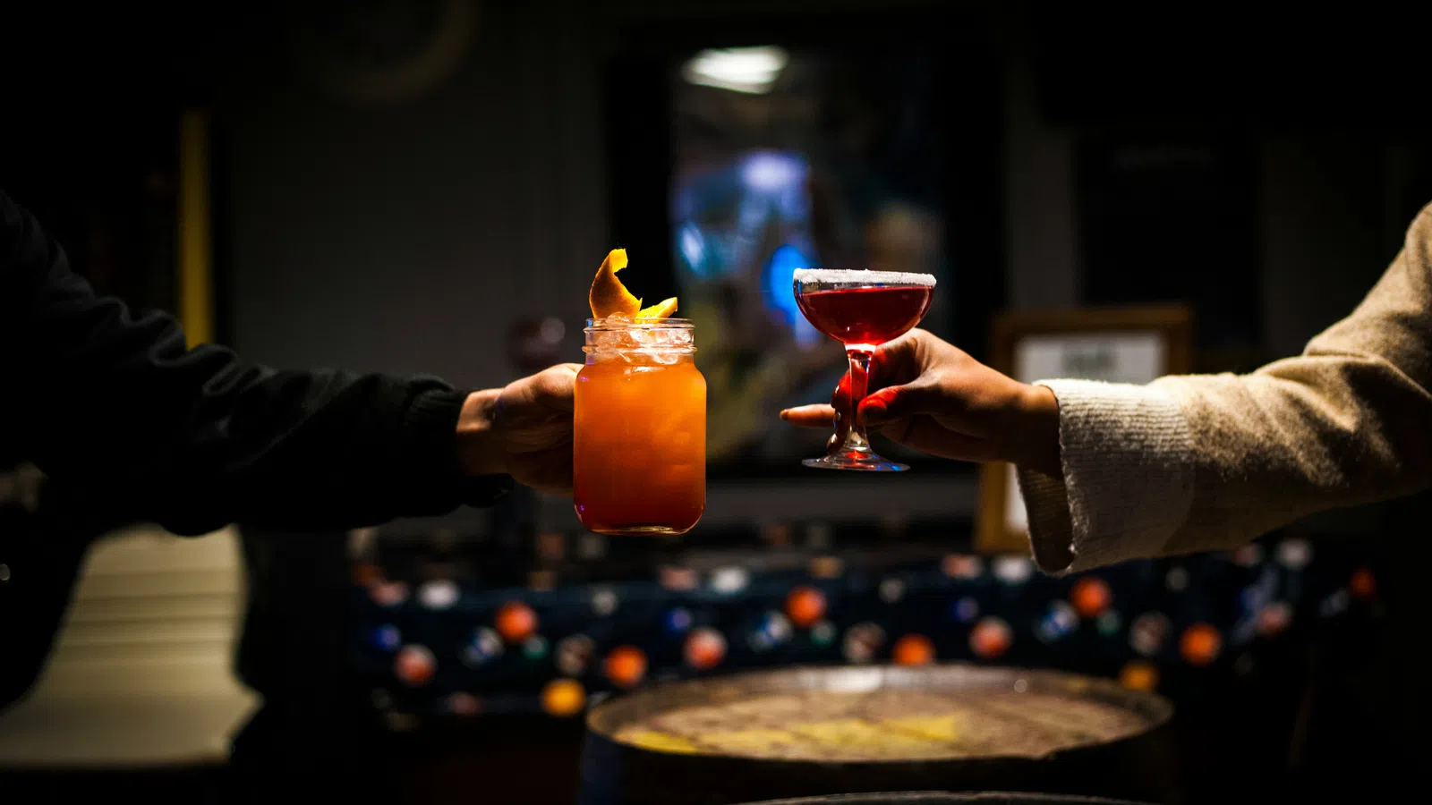 Two hands holding up colorful cocktails in dimly lit bar with wooden barrels in the foreground.