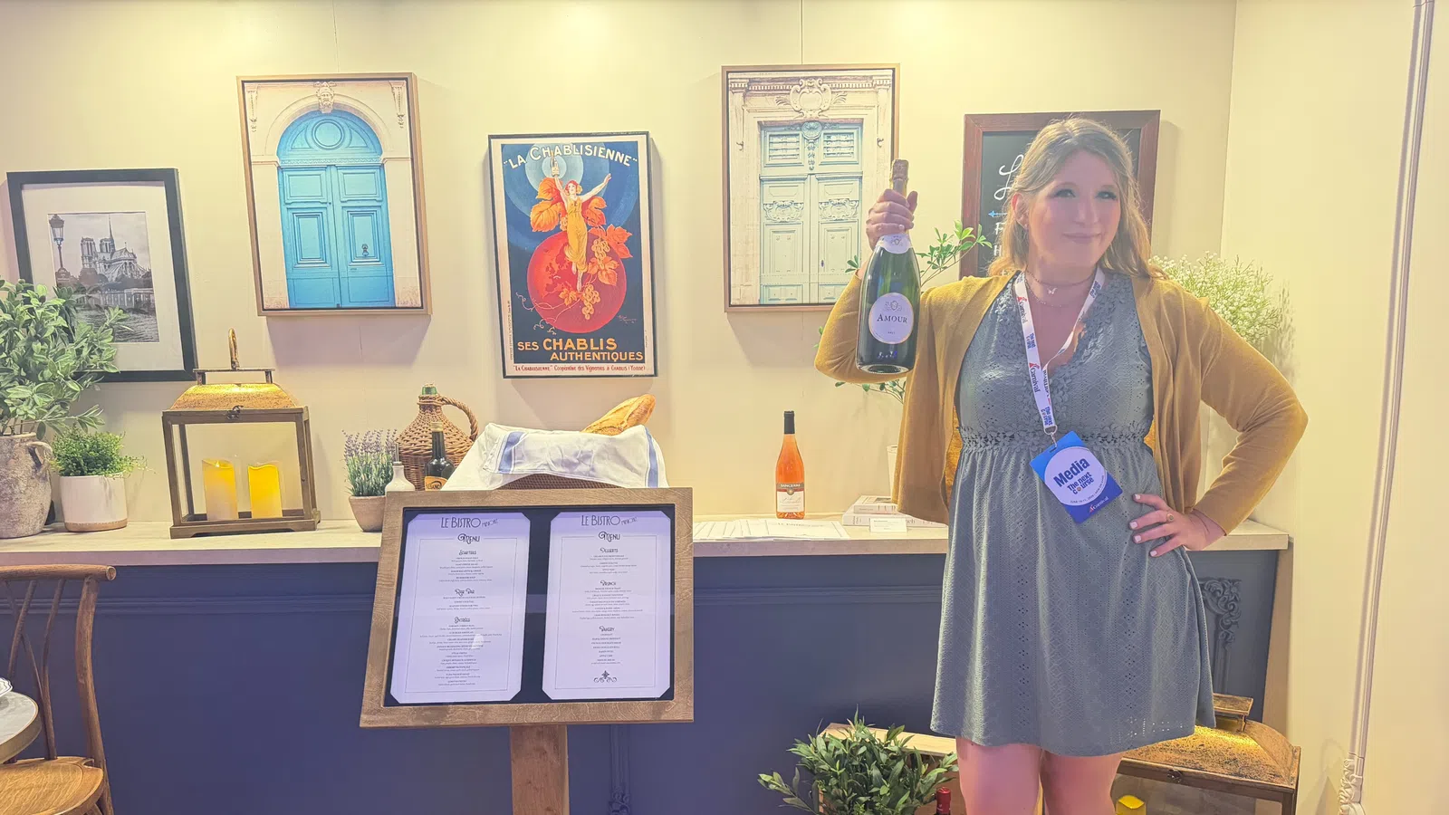 Woman holding wine bottle inside cozy wine bar with framed art, menu stand, plants, and candles in background.