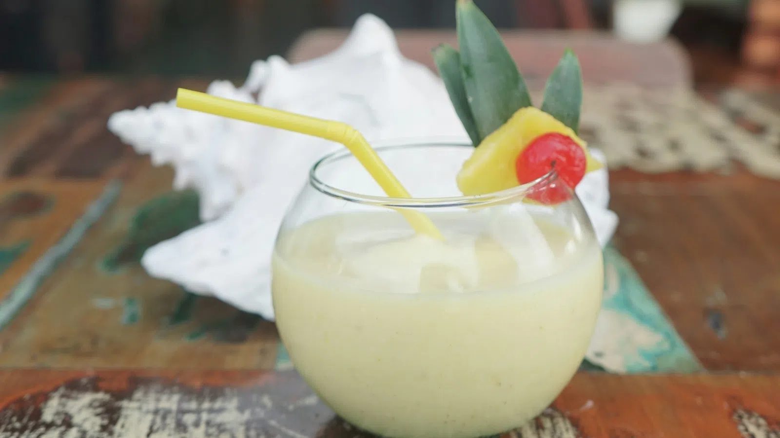 Pineapple smoothie in a round glass with a yellow straw, garnished with pineapple slice and cherry on a rustic wooden table.