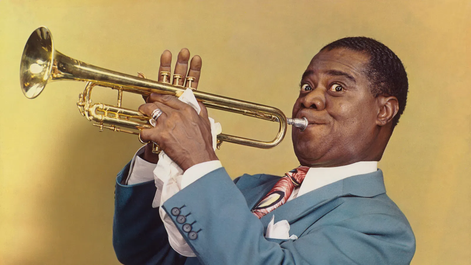 Man in blue suit playing trumpet with focused expression on yellow background.