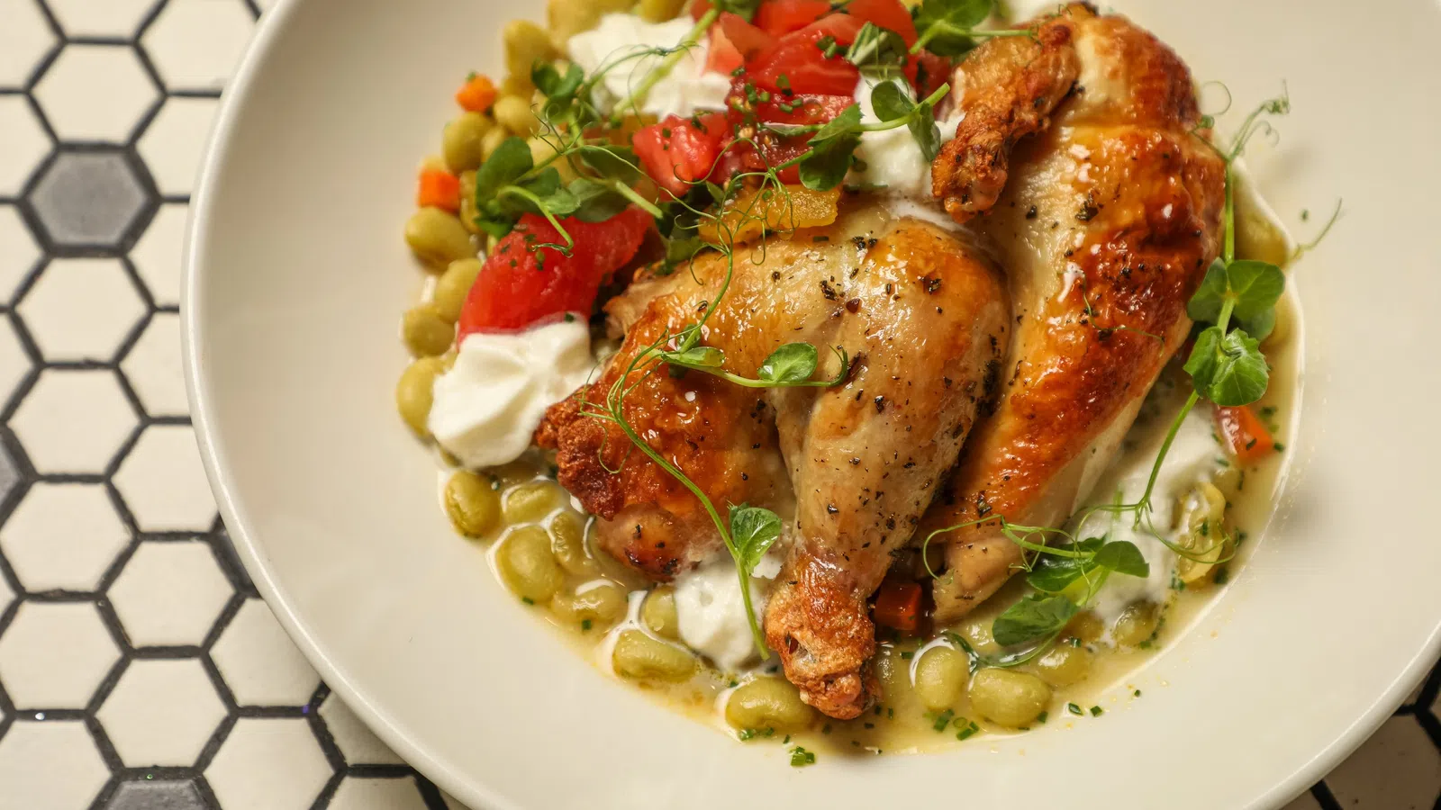 Roasted chicken leg quarters with white sauce, fava beans, diced tomatoes, and microgreens on white plate.
