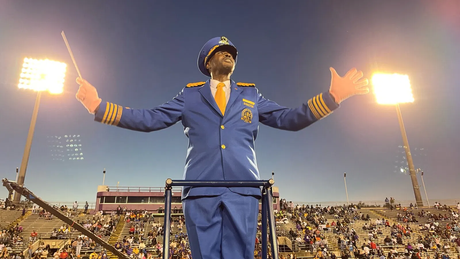 Band director in blue uniform conducting with baton at a stadium filled with spectators during evening.