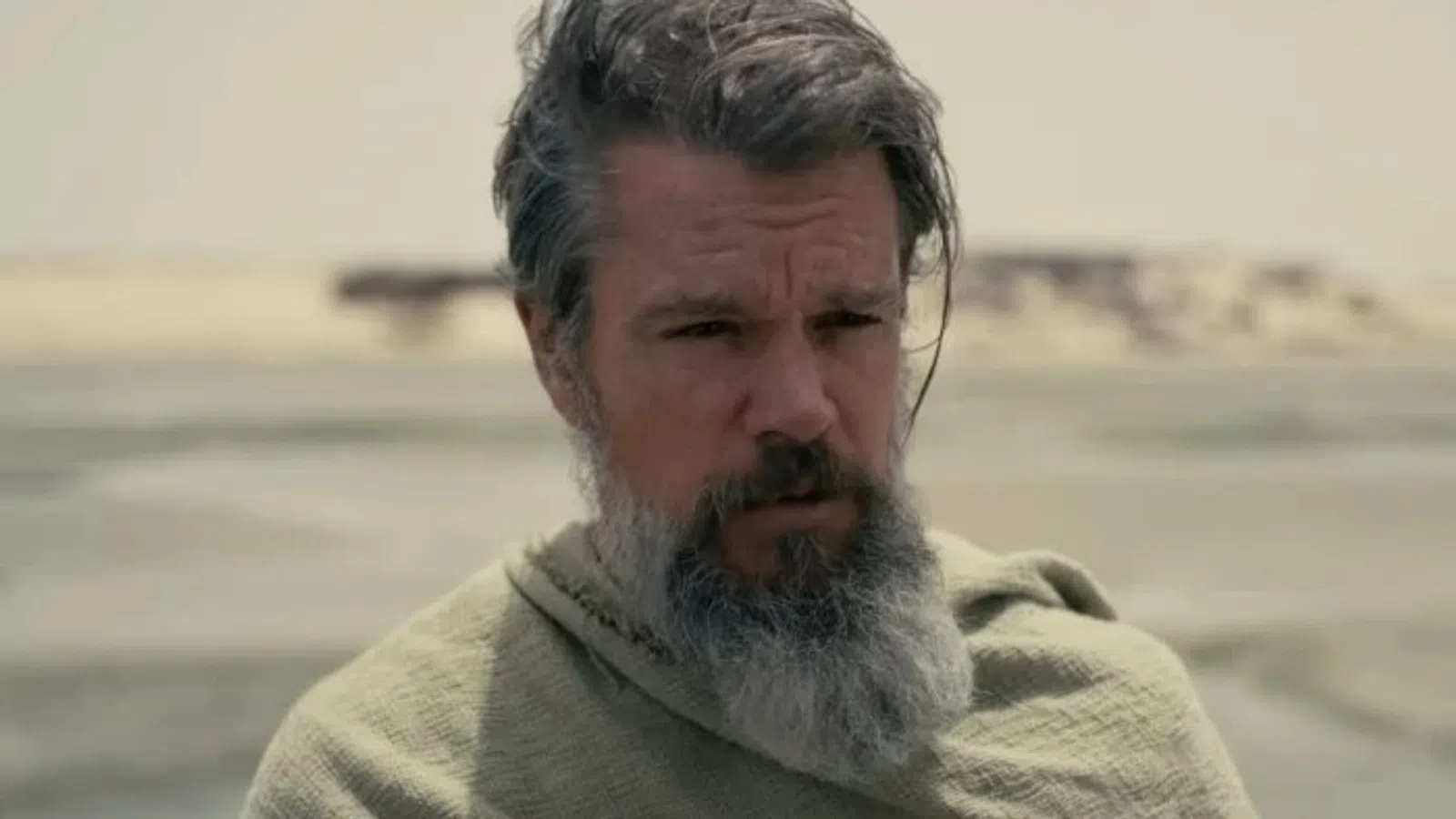 Man with gray beard and hair wearing a green cloak in a desert landscape with blurred sandy background.