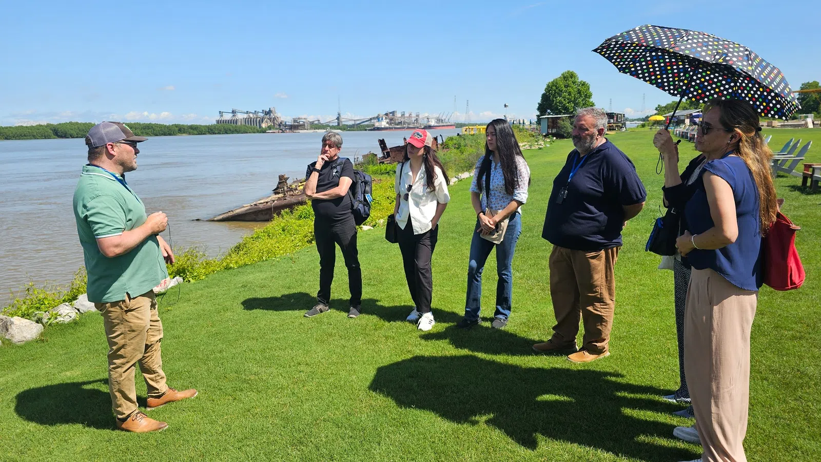 Group of six adults listening to a man speaking by a riverside on a sunny day with clear blue sky.