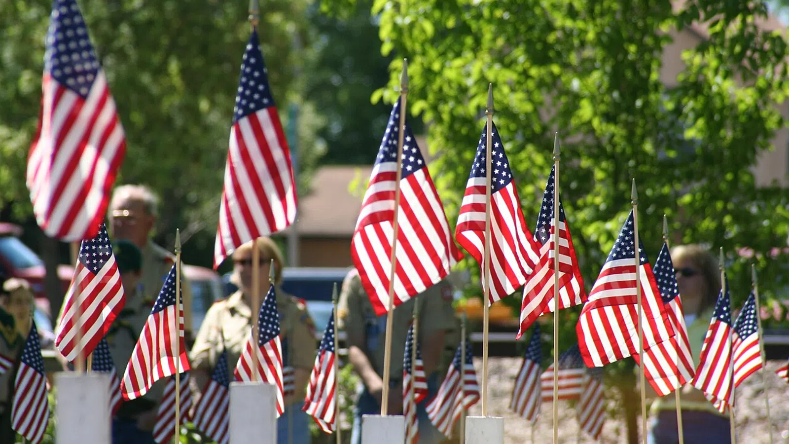 Multiple American flags on white posts with people blurred in the background on a sunny day