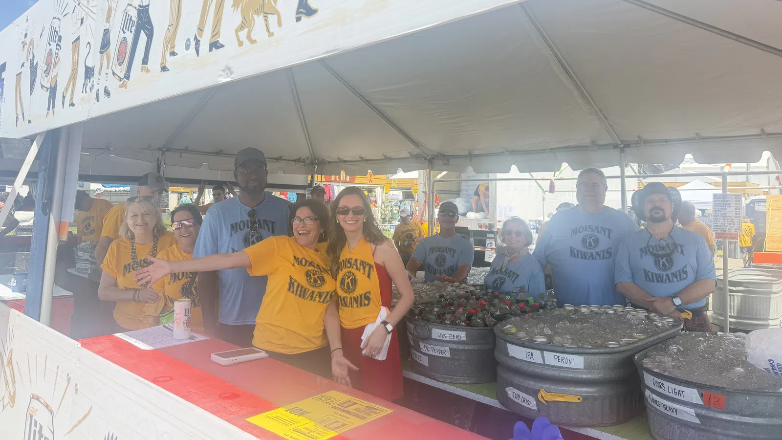 Group of Molsaint Kiwanis volunteers smiling and serving drinks under a festival tent with beer tubs.