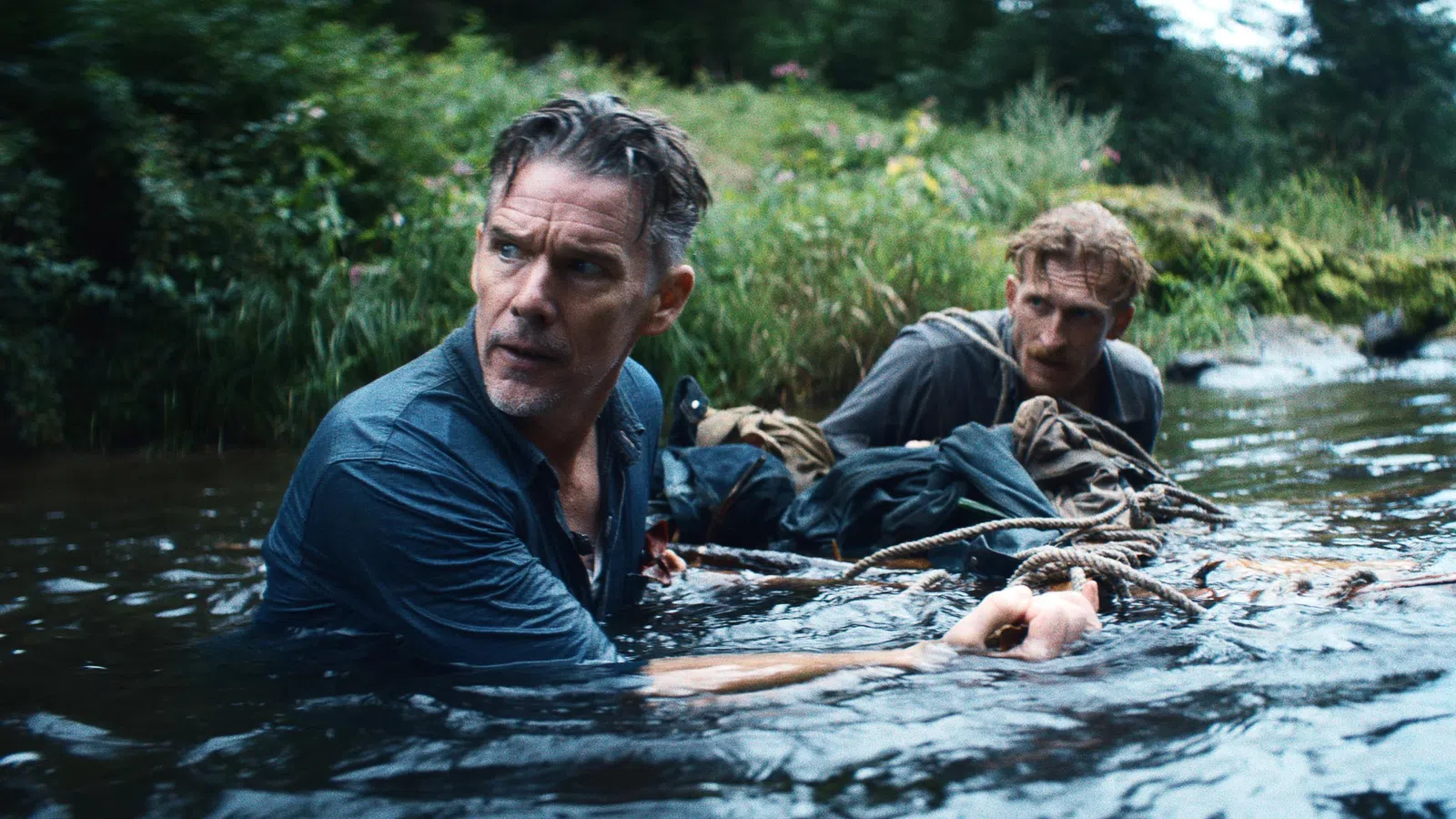 Two men cautiously wading through a river surrounded by dense greenery, holding onto a rope for support.