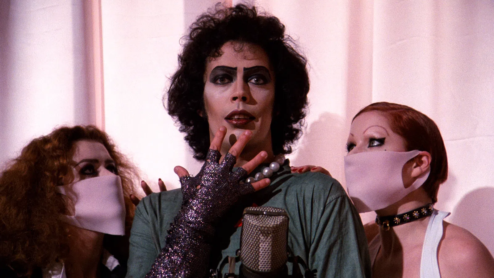 Actor in iconic glam rock makeup and costume with two masked fans on either side against a pale curtain.
