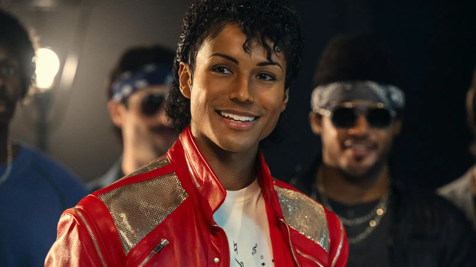 Smiling person wearing a red jacket with silver accents, with two others blurred in the background in dim lighting.