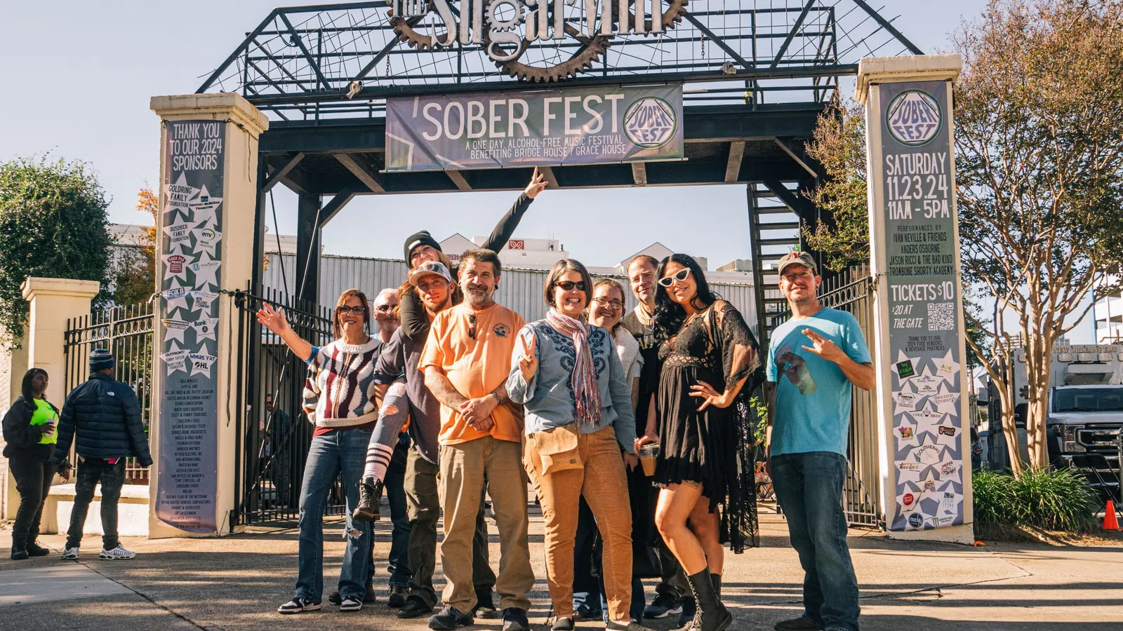 Group of people posing and smiling under Sober Fest banner at outdoor event entrance on sunny day
