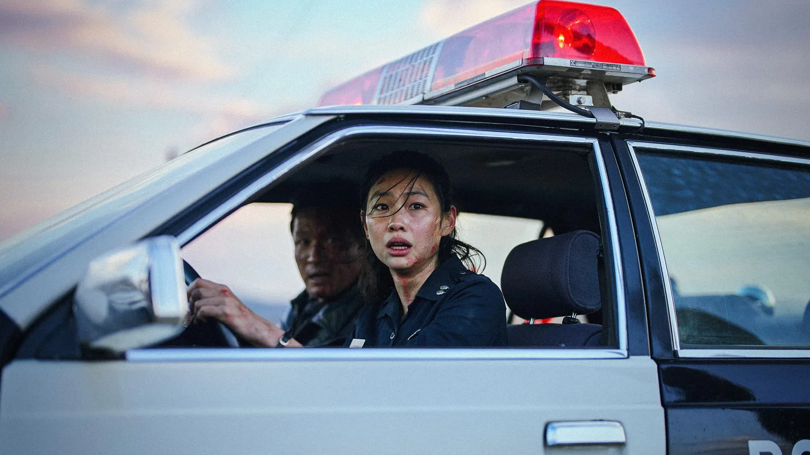 Female police officer and male colleague in a police car with red siren light, looking concerned through open window.